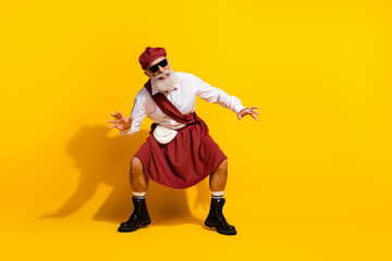 Elderly man in Scottish tartan outfit posing joyfully in yellow backdrop with stylish gestures, emphasizing fun and tradition