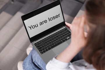 Cyber bullying. Upset teenage girl looking at laptop with text You Loser on stairs indoors, closeup