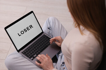 Cyber bullying. Upset teenage girl looking at laptop with text You Loser on floor indoors, closeup
