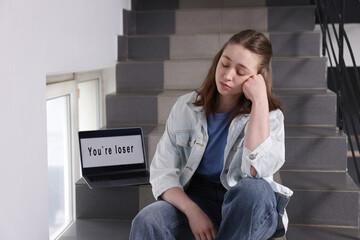 Cyber bullying. Upset teenage girl and laptop with text You Loser on stairs indoors