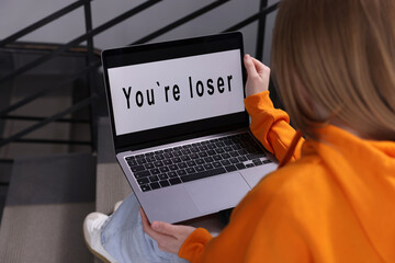 Cyber bullying. Upset teenage girl looking at laptop with text You Loser on stairs indoors, closeup