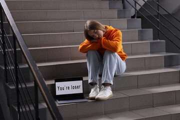 Cyber bullying. Upset teenage girl and laptop with text You Loser on stairs indoors