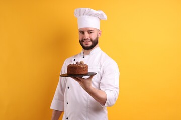 Happy confectioner in uniform holding chocolate cake on orange background