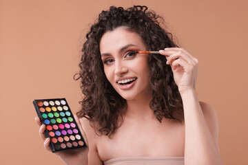 Smiling woman with eyeshadow palette and brush applying makeup on beige background