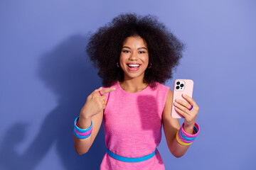 Joyful young woman in pink dress using smartphone with vibrant purple background, emphasizing...