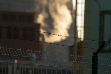 A plume of smoke is billowing out from a chimney situated just behind a barbed wire fence, creating...