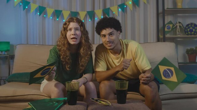 Multiracial couple celebrating a goal with a joyful kiss during a World Cup soccer match at home. Redhead woman and her partner wear Brazil colors and show passion, love and national pride.
