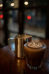 Close up of glass of hot latte with froth art and a jar on wooden table with bokeh background