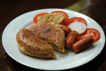 Canned Tuna-Filled Burger Toast comes with a Side Dish of Grilled Pork Sausage, Omelet Sliced Fresh Onion and Tomato in White Plate.