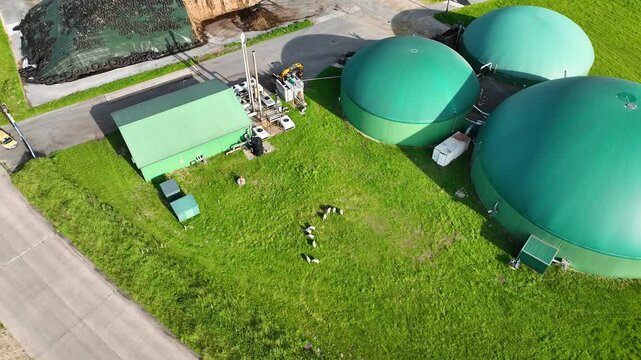 Aerial footage with camera tilt down to reveal green-roofed biogas digester facility, combined heat and power station with gas pipelines, technical units, silage storage. Sustainable energy production
