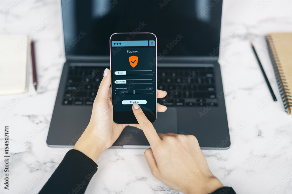 Poster person using smartphone to make secure online payment with laptop on desk in bright workspace backgr