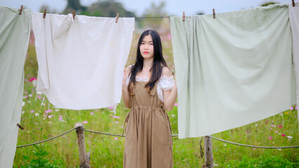 asian woman standing quietly among hanging fabrics in a field of flowers, wearing a brown dress and...