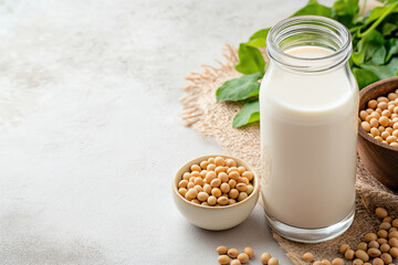 Glass bottle of soy milk sits on a textured surface beside bowls of soybeans and fresh green leaves