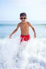 A joyful boy is playing in the water at the beach, splashing as waves crash around him. He wears sunglasses and bright red swim trunks, with a sunny sky above. Summer holidays concept. Generative AI.