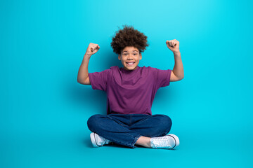 Cheerful young boy sitting cross-legged with vibrant gestures on a blue background showcasing happiness and confidence