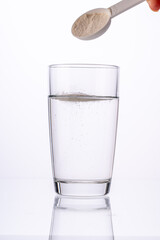 Collagen powder is poured with a measuring spoon into a glass of water on a white background with a reflection. The medicine is diluted in water. Vertical photo