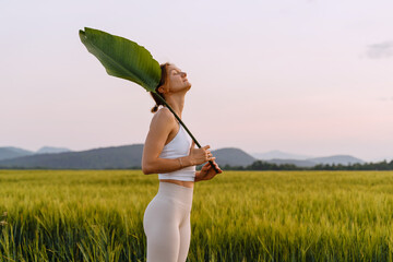 Portrait of relaxed and confident woman with big green leaf standing in middle in meadow.