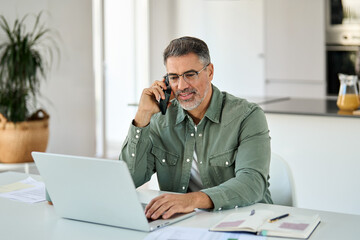 Happy busy middle aged mature man senior entrepreneur talking on mobile phone looking at laptop using computer calling on cellphone working at home sitting at kitchen table.