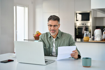 Middle aged mature man senior customer wearing glasses looking at laptop computer holding paper bill checking financial invoice or tax document making online banking payments working at home table.