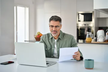  Middle aged mature man senior customer wearing glasses looking at laptop computer holding paper bill checking financial invoice or tax document making online banking payments working at home table. © insta_photos