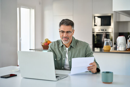 Fototapeta Middle aged mature man senior customer wearing glasses looking at laptop computer holding paper bill checking financial invoice or tax document making online banking payments working at home table.