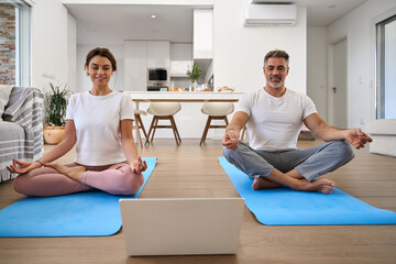 Happy middle aged family couple doing yoga at home looking at laptop. Healthy calm serene mature older man and woman doing exercises meditating watching wellness tutorial in living room.