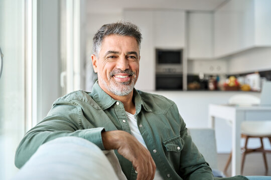 Close up portrait of happy smiling middle aged mature senior 50 years old bearded man wearing green shirt sitting on couch at home interior looking at camera relaxing on sofa in modern house.