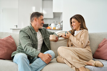 Happy middle aged mature couple talking relaxing on couch at home. Relaxed smiling older man and woman in love sitting on sofa together enjoying tea and conversation in modern house living room.