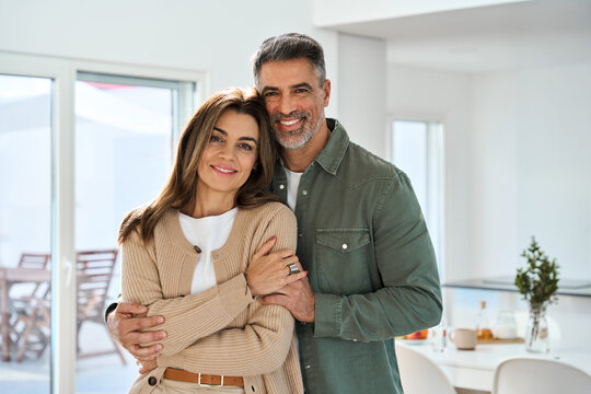 Fototapeta Smiling affectionate loving middle aged couple, happy romantic mature older Latin man and woman in their 50s hugging looking at camera standing together in modern kitchen at home. Portrait.