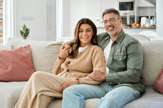 Family portrait of happy middle aged mature romantic couple in their 50s hugging sitting on couch at home. Smiling older man and woman in love relaxing on sofa together in house living room.