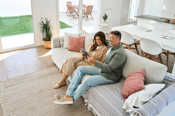 Happy mature old couple middle aged man and woman hugging sitting on sofa at home relaxing on couch using digital tablet buying online browsing in modern house living room. Top view from above.