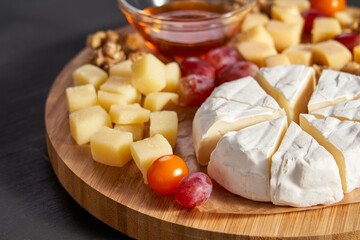 A wooden tray with a variety of cheeses and fruit, including grapes and oranges. The cheese is cut...