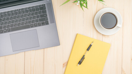 A laptop and a cup of coffee sit on a wooden table. The laptop is open and the keyboard is visible. The cup of coffee is placed next to the laptop, and a pen is resting on the yellow notebook