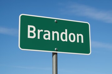 Green road sign with "Brandon" in white text, standing beneath a clear blue sky with thin clouds, marking city entry.