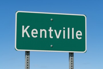 Green road sign displaying "Kentville" beneath a bright blue sky, indicating town entrance, travel direction, or Nova Scotia destination.