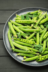 Fresh green peas in plate with pods and leaves on black wooden background. Vertical photo