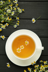 Aromatic herbal tea in white cup and chamomiles on black wooden background. Vertical photo