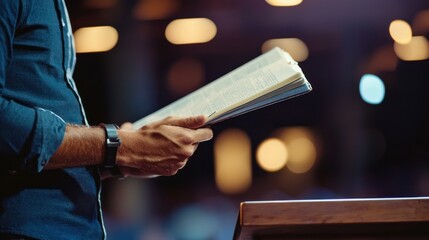 A man in a blue shirt reads from a book on a podium in a dimly lit room with a blurred background.