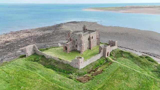 Aerial drone orbit of Piel Castle ruins on the coast of Cumbria, England. The historic stone structure sits isolated on the edge of the sea, surrounded by grassland and revealed shoreline at low tide.