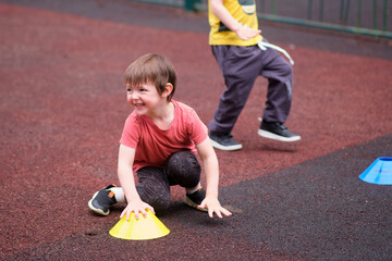 Smiling children enjoy active play with colorful cones on a rubber playground surface during an outdoor group activity. A child aged 3 years