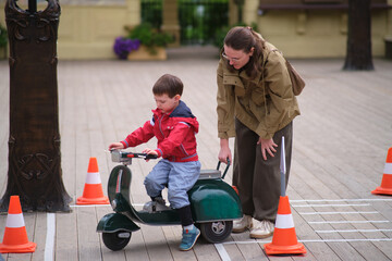 Woman helping a boy to ride a toy scooter. Kid learns to balance a scooter on a wooden path. Childhood education and parenting. A child aged 3 years