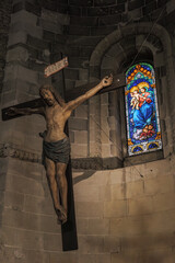 Matera, Italy. Wooden crucifix of Jesus in the Church of San Giovanni Battista, Sassi of Matera. A stained glass window depicting the Madonna and Child glows in the background. Vertical. 