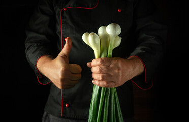 In a dimly lit kitchen, a chef proudly holds a bunch of fresh green onions while giving a thumbs up. The atmosphere conveys enthusiasm for cooking with fresh ingredients