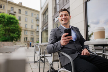 Smiling man in a suit enjoys sunny day at outdoor cafe while checking messages on smartphone in urban setting
