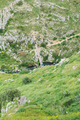 Matera, Italy. Scenic view of the Gravina river below Matera's Sassi. People cross a Tibetan bridge along path 406 in the Murgia Materana Park, surrounded by rocky green hills. Vertical image.