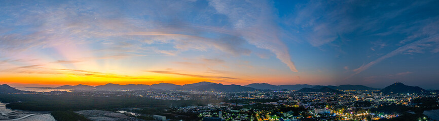 A stunning panoramic view of a coastal city at sunset at Phuket city, showcasing vibrant hues of orange and blue in the sky, with mountains, and urban lights creating a harmonious blend of natural