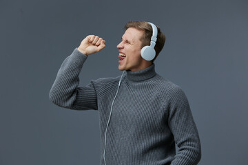 Happy young man wearing gray turtleneck sweater enjoying music with large white headphones on plain gray background, celebrating, feeling cheerful, relaxed, casual style, lifestyle concept