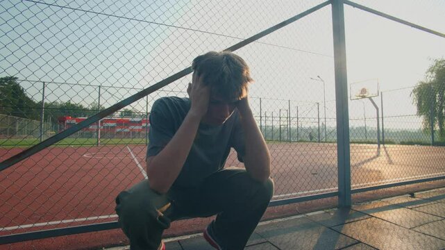 Teenager sits near sports field with hands on face in emotional moment
