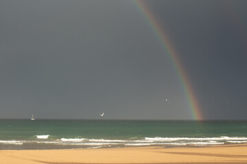 Beautiful rainbow arches over the ocean as sailboats glide peacefully along the Spanish coastline