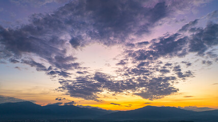 A breathtaking aerial panoramic view of a vibrant sunset illuminating the sky above a lush green hilltop with a small structure, surrounded by scattered clouds and distant landscapes.